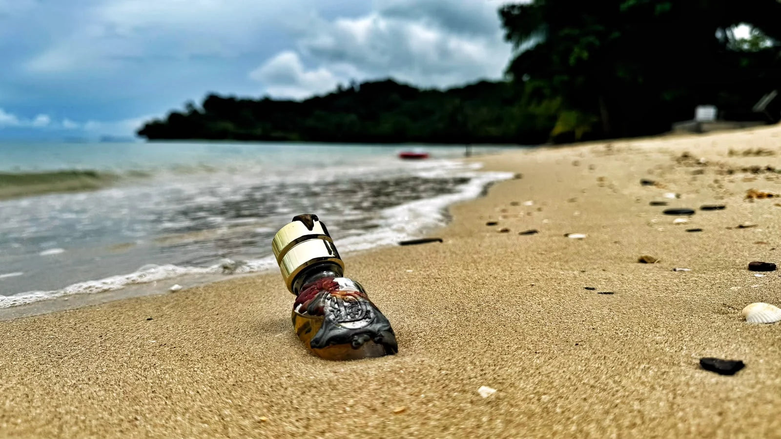 Ein Flakon mit Wachssiegel liegt im hellen Sand eines thailändischen Strandes, links laufen die Wellen aus, im Hintergrund eine bewaldete Bucht
